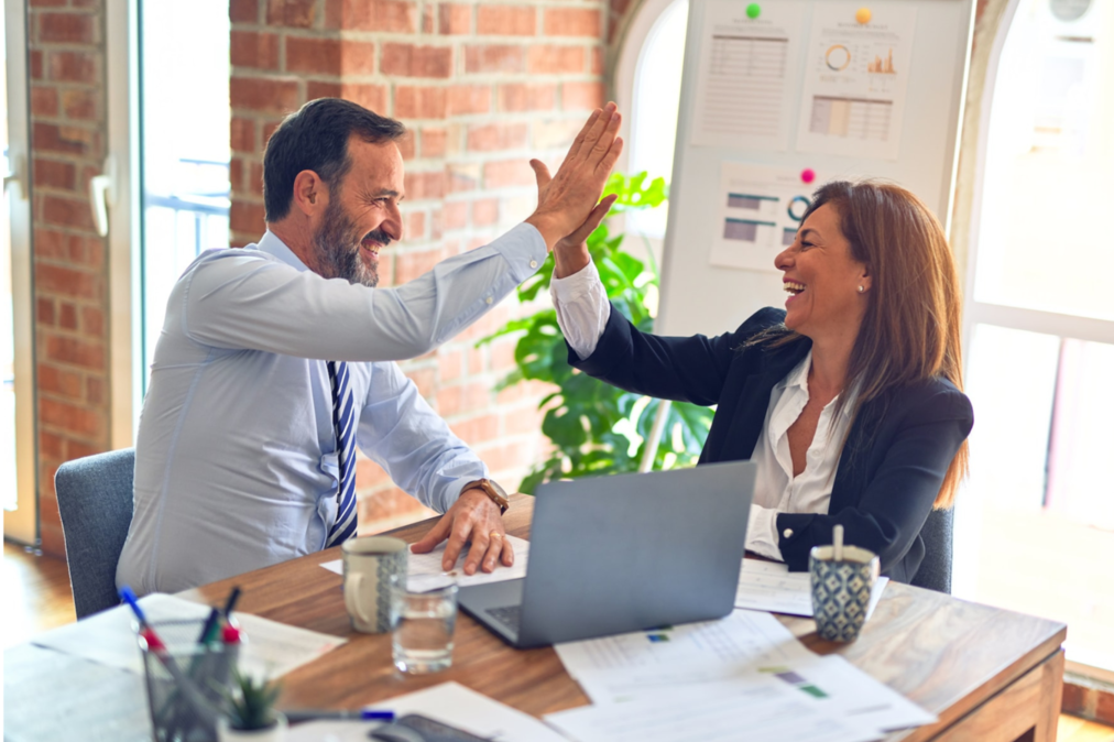 Two people high fiving at a desk in an office in front of a computer